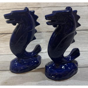 May include: Two cobalt blue ceramic seahorse figurines. Each seahorse has a detailed head, spiky dorsal fin, and a curled tail. The figurines are mounted on a textured base, creating a decorative nautical theme.