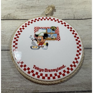 May include: A round ceramic wall plaque featuring a red and white checkered border and a rope hanger. The center displays Mickey Mouse as a chef cooking eggs, with a window view of a castle and the text Tokyo Disneyland printed in red.
