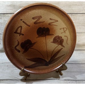 Puede incluir: Un plato redondo de cerámica marrón con un diseño floral y la palabra "Pizza" escrita alrededor del borde. El plato está sostenido por un soporte de metal dorado. El plato tiene un aspecto rústico y hecho a mano.