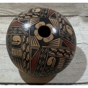 Puede incluir: Un recipiente de cerámica redondo, hecho a mano, con un agujero central y patrones geométricos intrincados. El diseño presenta formas negras, beige y marrón rojizo, incluyendo triángulos, líneas y figuras de pájaros estilizadas. El recipiente probablemente está pintado a mano.