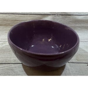 Peut inclure: Un bol en céramique violet foncé avec une finition brillante. Le bol a une forme arrondie et un intérieur lisse. L'extérieur a un aspect texturé. Le bol est posé sur une surface en bois de couleur claire.