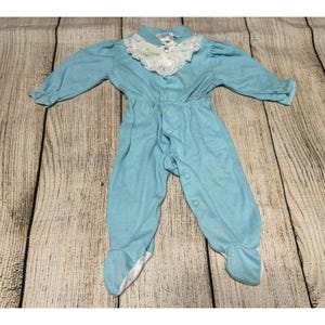 May include: A light blue baby onesie with long sleeves and attached feet. The onesie has a white lace-trimmed collar with a floral pattern. It fastens with buttons down the front.