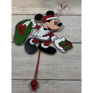 May include: Wooden Christmas ornament featuring Mickey Mouse dressed as Santa Claus. Mickey is holding a green wreath with a red bow and a wrapped gift. The ornament is painted in red, black, white, and tan colors.
