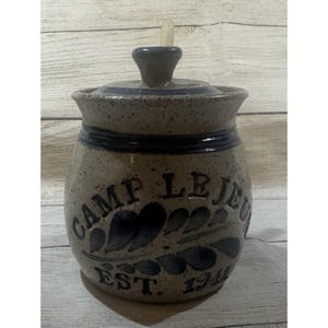 May include: A speckled beige ceramic crock with a lid and a handle. The front features dark blue painted text that reads CAMP LEJEUNE and EST. 1942, accompanied by a dark blue leaf-like decorative pattern. The crock has a wide rim and a rounded body.