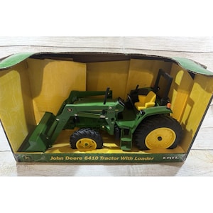 Puede incluir: Un tractor John Deere 6410 verde y amarillo con cargador, exhibido en su caja original. El tractor tiene neumáticos negros con llantas amarillas y un asiento amarillo. La caja tiene una combinación de colores verde y amarillo.