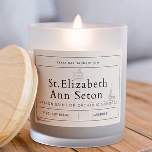 May include: A lit, frosted glass candle with a wooden lid. The label reads "St. Elizabeth Ann Seton, Patron Saint of Catholic Schools." The candle is 11 oz and soy blend, with "Lavender" scent. The text "Feast Day January 4th" is also visible.