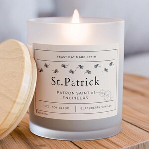 May include: A lit, frosted glass candle with a wooden lid. The label reads "St. Patrick, Patron Saint of Engineers" and "11 oz Soy Blend Blackberry Vanilla." The candle is on a wooden surface.