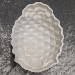 Könnte beinhalten: Eine weiße, ananasförmige Servierschale. Die Schale hat eine strukturierte Oberfläche mit einem geometrischen Muster und eine detaillierte Oberseite, die einer Ananaskrone ähnelt. Die Schale besteht aus Keramik oder Porzellan.