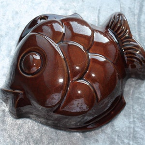 Può includere: Uno stampo in ceramica marrone scuro lucido a forma di pesce. Lo stampo presenta una superficie liscia e riflettente con dettagli modellati che rappresentano l'occhio, le squame e le pinne caudali del pesce. Il design è di forma concava.