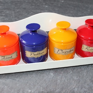 Peut inclure: Un présentoir à épices en bois blanc avec quatre pots à épices en céramique colorés. Les pots sont orange, bleu, jaune et rouge, chacun avec un couvercle assorti. Chaque pot a une étiquette avec un nom d'épice.