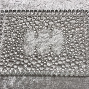 May include: A rectangular, clear glass tray with a bubbly, textured surface. The tray's border is lined with small, rounded glass beads. The interior features a design of larger, bubble-like shapes, creating a decorative effect.