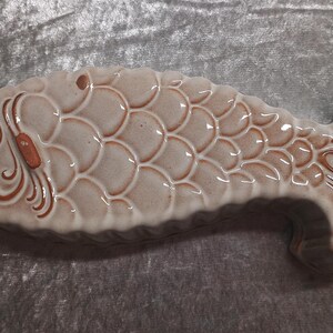 May include: A ceramic fish-shaped dish with a glossy finish. The dish is primarily beige with brown accents, featuring a detailed scale pattern and decorative elements on the head and tail. The fish measures approximately 12 inches in length.