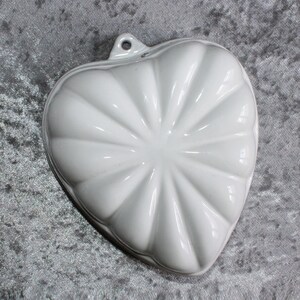 Può includere: Stampo a forma di cuore in ceramica bianca con un design a coste. Lo stampo ha un piccolo manico ad anello nella parte superiore.