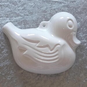 Op de afbeelding: Een witte keramische eendenfiguur met een glanzende afwerking. De eend heeft gedetailleerde vleugel- en veermotieven, een klein oog en een lus om op te hangen. Het beeldje is een decoratief item, mogelijk voor een kinderkamer of keuken.