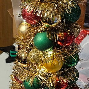 Op de afbeelding: Een kleine gouden kerstboom versierd met rode, groene en gouden kerstballen. De boom is bedekt met gouden tinsel en heeft een gouden ster bovenop.