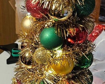 May include: A small gold Christmas tree decorated with red, green and gold baubles. The tree is covered in gold tinsel and has a gold star on top.