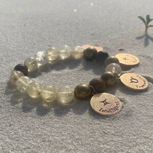 Könnte beinhalten: Ein goldenes Armband mit klaren und braunen Perlen und drei goldenen Anhängern mit Sternzeichen. Die Anhänger sind mit den Worten "Zwillinge, Intelligent, Vertrauenswürdig" graviert.