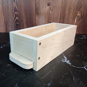 May include: A rectangular wooden soap mold with a removable bottom. The mold is made of light wood and has a smooth finish. It is designed for making homemade soap.