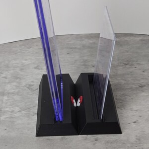 May include: A black, two-part display stand with clear, upright panels. The panels have a blue tint and are held in place by the stand. A small, red and white butterfly-shaped accent is between the panels.
