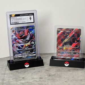May include: Two Pokemon trading cards in protective cases. One card features a red and black character with Japanese text, graded as a 9. The other card shows a red and black character with the text "Burning Slam GX". Both cards are displayed on black stands with a Pokemon logo.