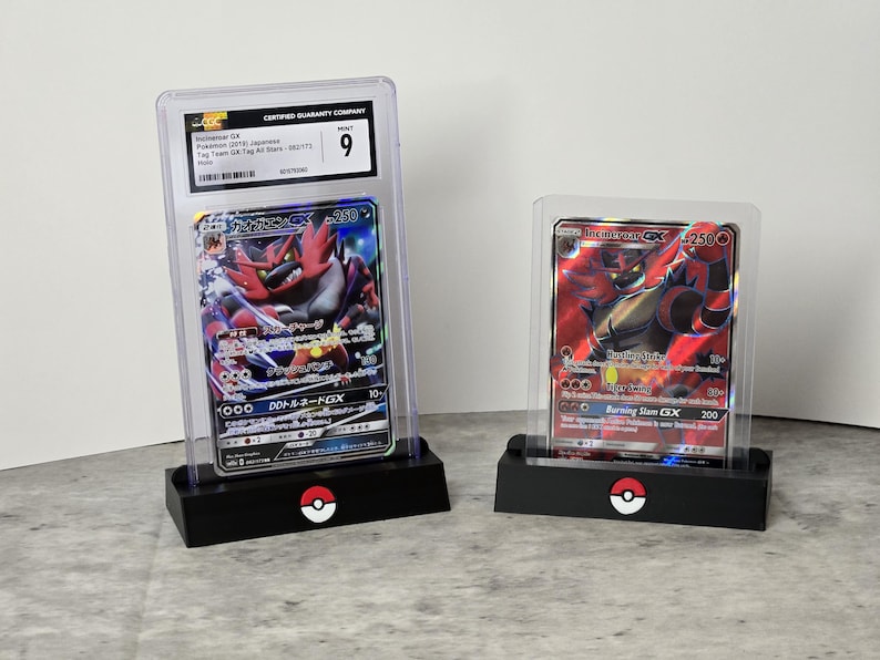 May include: Two Pokemon trading cards in protective cases. One card features a red and black character with Japanese text, graded as a 9. The other card shows a red and black character with the text "Burning Slam GX". Both cards are displayed on black stands with a Pokemon logo.