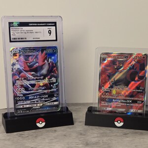 May include: Two Pokemon trading cards in protective cases. One card features Incineroar GX with Japanese text and a CGC grade of 9. The other card also features Incineroar GX. Both cards are displayed on black stands with a Pokemon logo.