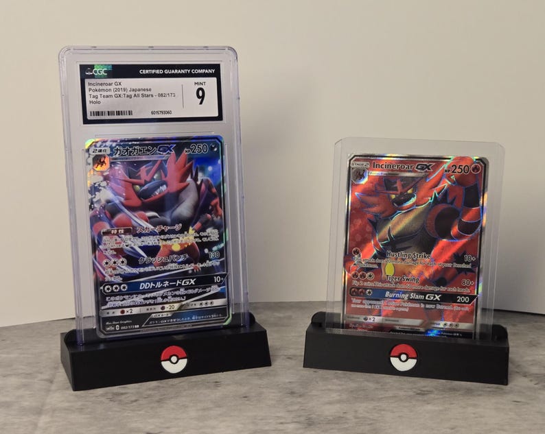 May include: Two Pokemon trading cards in protective cases. One card features Incineroar GX with Japanese text and a CGC grade of 9. The other card also features Incineroar GX. Both cards are displayed on black stands with a Pokemon logo.