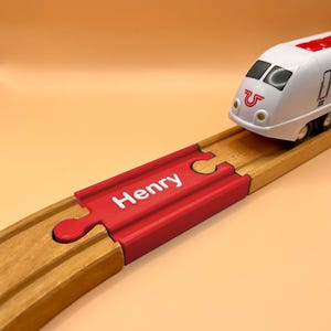 May include: A white toy train engine with black windows and red accents sits on a wooden train track. A red track connector piece is inserted into the wooden track, featuring the name Henry printed in white letters on its top surface.