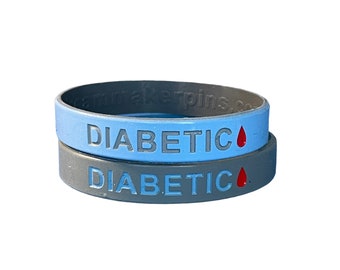 Puede incluir: Dos pulseras de silicona, una azul y otra gris, con la palabra "DIABÉTICO" y una gota de sangre roja impresa en cada una.