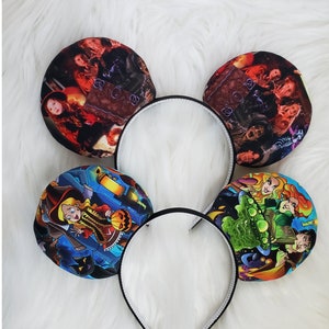 May include: Four black headbands with circular ear-shaped decorations. The decorations feature colorful illustrations of characters and scenes, likely from a movie or cartoon. The headbands are on a white, fluffy surface.