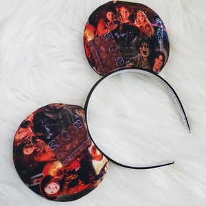 May include: A pair of black headband ears featuring a colorful image of characters from the movie "Hocus Pocus". The ears are round and attached to a black headband with a white, textured interior.