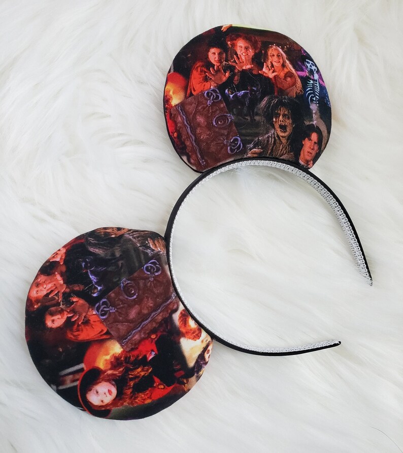May include: A pair of black headband ears featuring a colorful image of characters from the movie "Hocus Pocus". The ears are round and attached to a black headband with a white, textured interior.