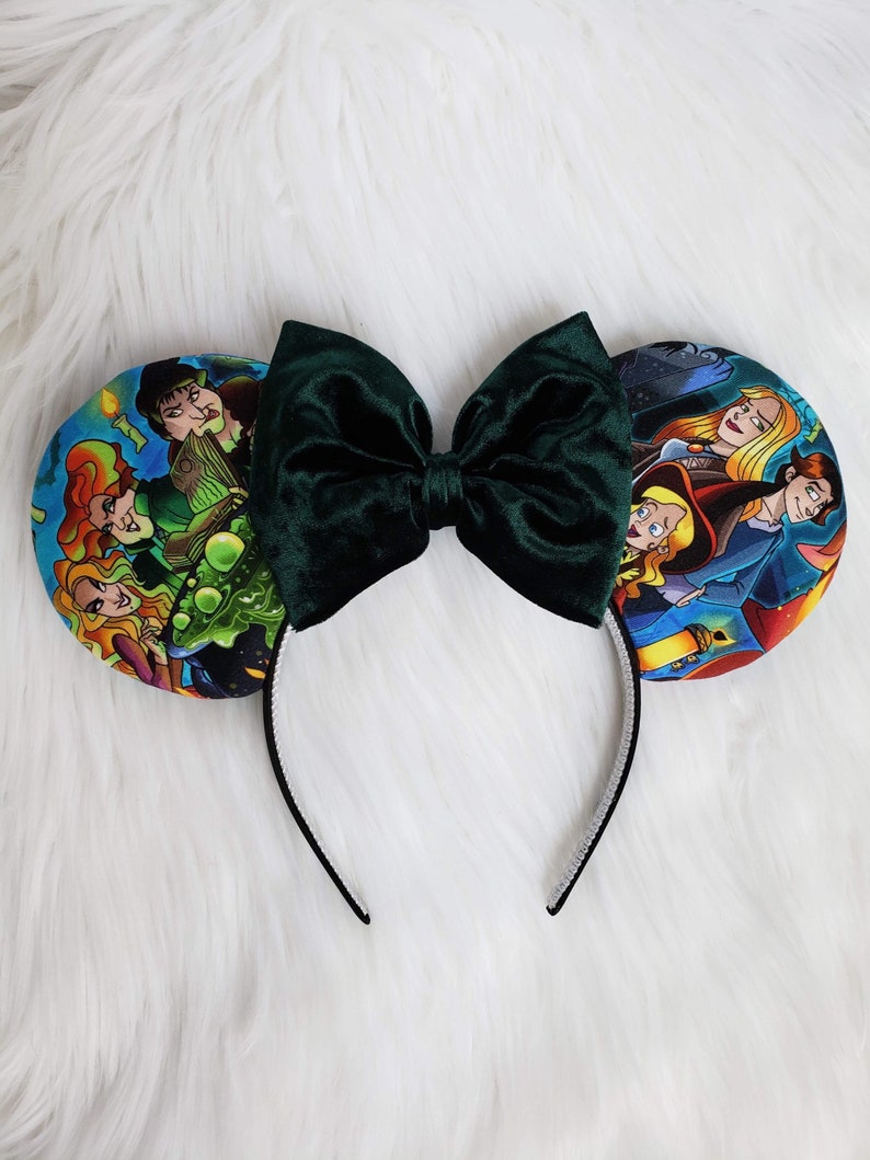 May include: A pair of mouse ears with a dark green velvet bow and character-themed ear designs. The ears feature colorful illustrations of characters on a white headband.
