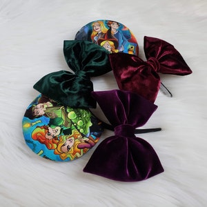 May include: Several velvet bow headbands in shades of green, burgundy, and purple. The bows are attached to black headbands. Two headbands feature colorful circular designs with cartoon characters.