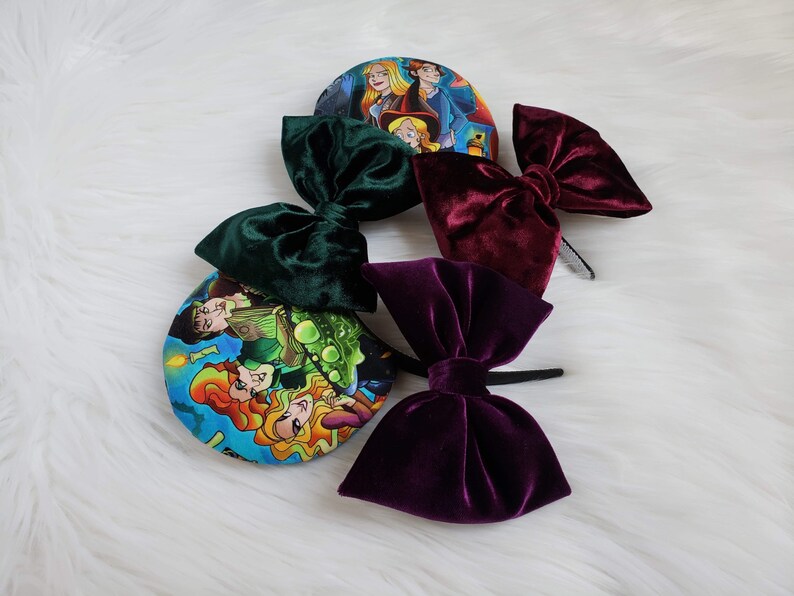 May include: Several velvet bow headbands in shades of green, burgundy, and purple. The bows are attached to black headbands. Two headbands feature colorful circular designs with cartoon characters.