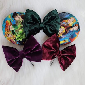 May include: An assortment of velvet hair bows in deep green, burgundy, and purple, alongside two round ear headbands with a colorful cartoon print. The bows are attached to black headbands.