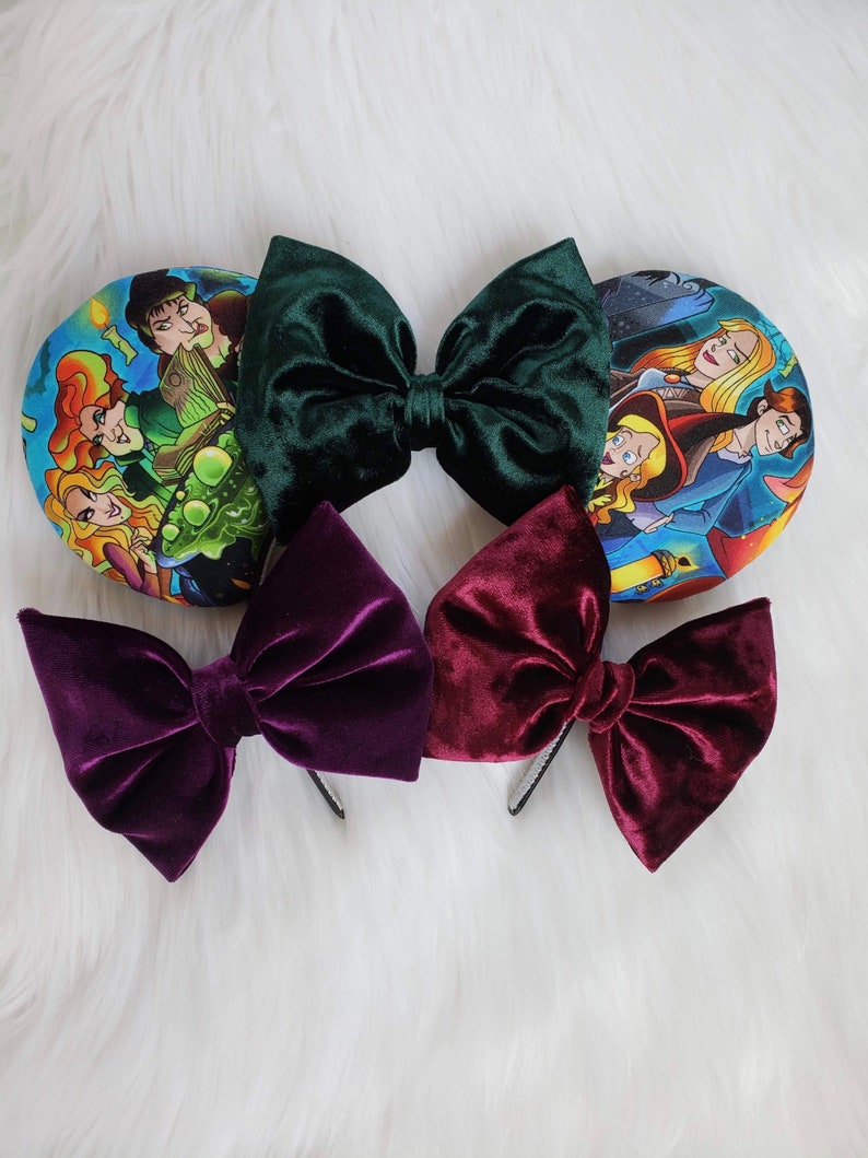 May include: An assortment of velvet hair bows in deep green, burgundy, and purple, alongside two round ear headbands with a colorful cartoon print. The bows are attached to black headbands.