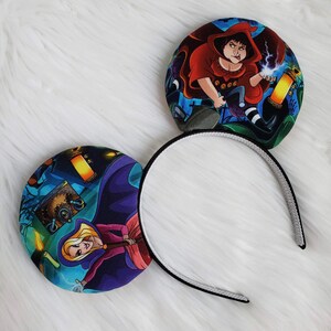 May include: A pair of mouse ear headbands featuring colorful illustrations of characters in a spooky setting. The ears are round with a black and white headband.