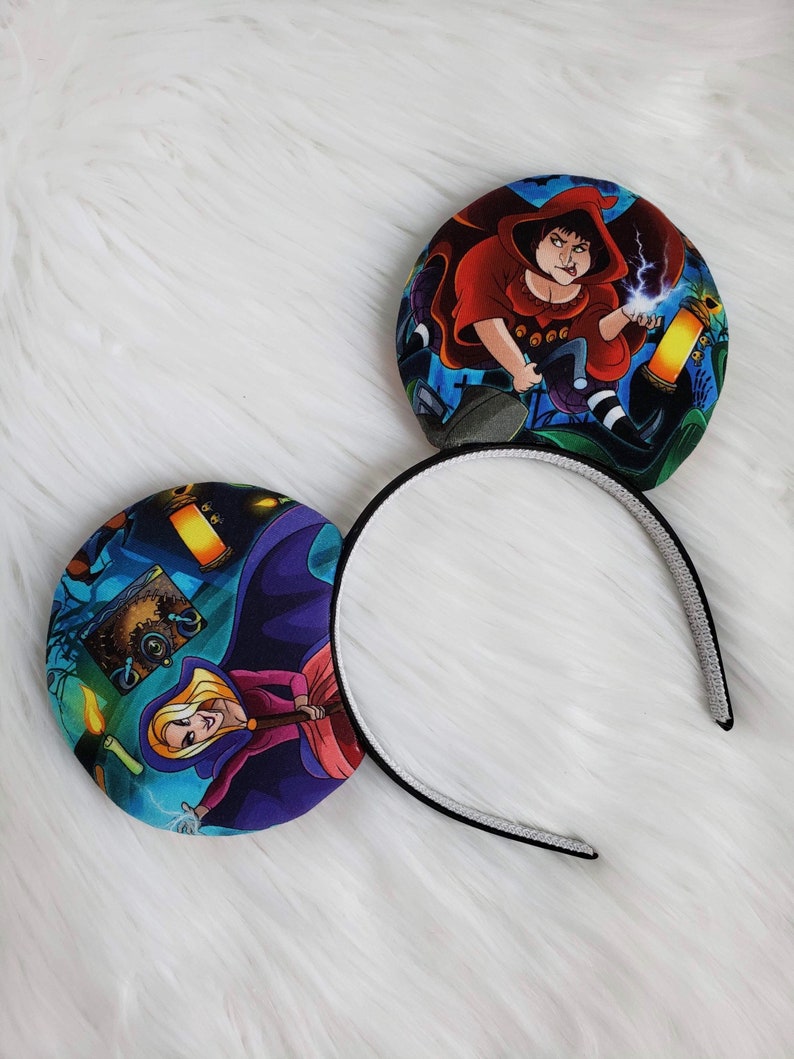 May include: A pair of mouse ear headbands featuring colorful illustrations of characters in a spooky setting. The ears are round with a black and white headband.