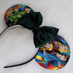 May include: A pair of themed mouse ears featuring a dark green velvet bow and character art on the ear pads. The headband is black with a silver rhinestone trim. The ear pads display colorful illustrations.