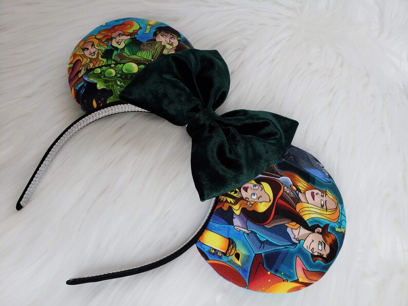 May include: A pair of themed mouse ears featuring a dark green velvet bow and character art on the ear pads. The headband is black with a silver rhinestone trim. The ear pads display colorful illustrations.