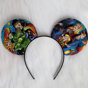 May include: A pair of mouse ears with a black headband and two circular ear designs. The ears feature colorful illustrations of characters from a fantasy movie. The headband is black with a white interior.