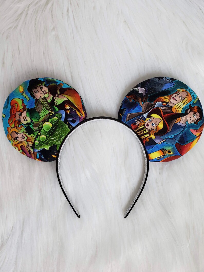 May include: A pair of mouse ears with a black headband and two circular ear designs. The ears feature colorful illustrations of characters from a fantasy movie. The headband is black with a white interior.