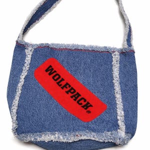 May include: Blue denim shoulder bag with a red rectangular patch that reads "WOLFPACK." The bag has a frayed edge design and a matching denim strap. The bag is made of blue denim material.