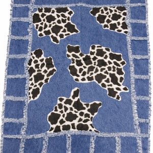 May include: Blue denim rug with a cow print design. The rug features a blue denim base with five black and white cow print shapes. The edges are trimmed with a frayed denim border.