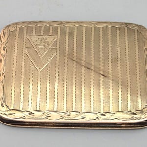 May include: A rectangular gold-colored box with a vertically striped pattern and a decorative border. A triangular emblem is centered on the lid. The box is likely vintage and may be used for storing small items.