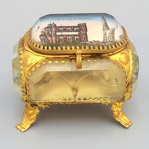 Puede incluir: Una pequeña caja de vidrio de color dorado con una tapa con bisagras. La tapa presenta una imagen pintada de un edificio con una torre en el fondo. La caja tiene tres patas y está decorada con un borde dorado.