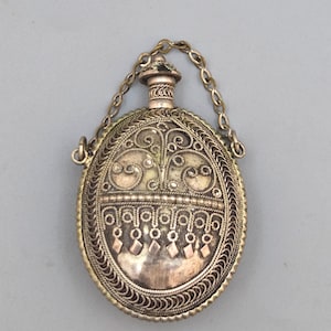 May include: An antique silver-toned perfume bottle with an oval filigree design. The bottle features intricate scrollwork, a decorative top, and a chain. The design includes diamond-shaped accents and a detailed border.