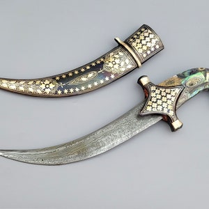 Puede incluir: Una daga decorativa con una hoja plateada y un mango negro y dorado. El mango está incrustado con nácar y diseños florales dorados. La daga viene con una vaina a juego.