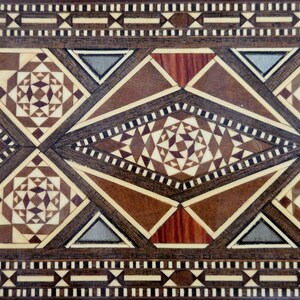 May include: Close-up of a wooden surface with intricate geometric patterns. The design features various shapes, including diamonds, triangles, and stars, in shades of brown, beige, and gray. The detailed craftsmanship suggests a decorative or ornamental item.
